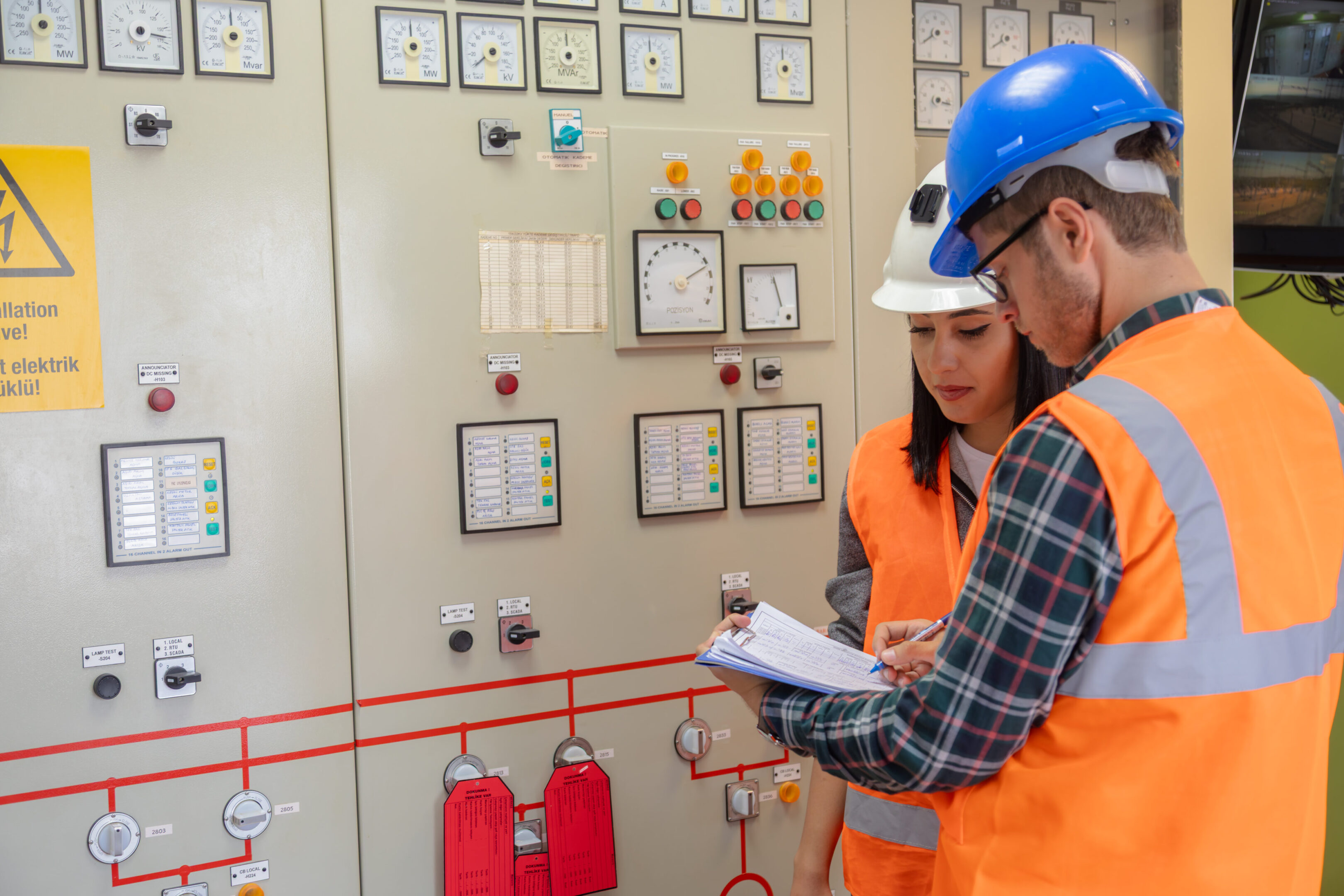 A male and female engineer, both wearing high-visibility vests and safety helmets, collaborate in the bright control room of a renewable energy facility. They review technical documents together in front of an advanced control panel with monitoring gauges, switches, and digital interfaces. In the background, other team members work and observe, emphasizing the importance of teamwork, detailed documentation, and occupational safety in energy management. The scene highlights modern engineering practices, digital solutions, and the integration of smart technology and artificial intelligence for efficient system monitoring, predictive maintenance, and sustainable energy operations